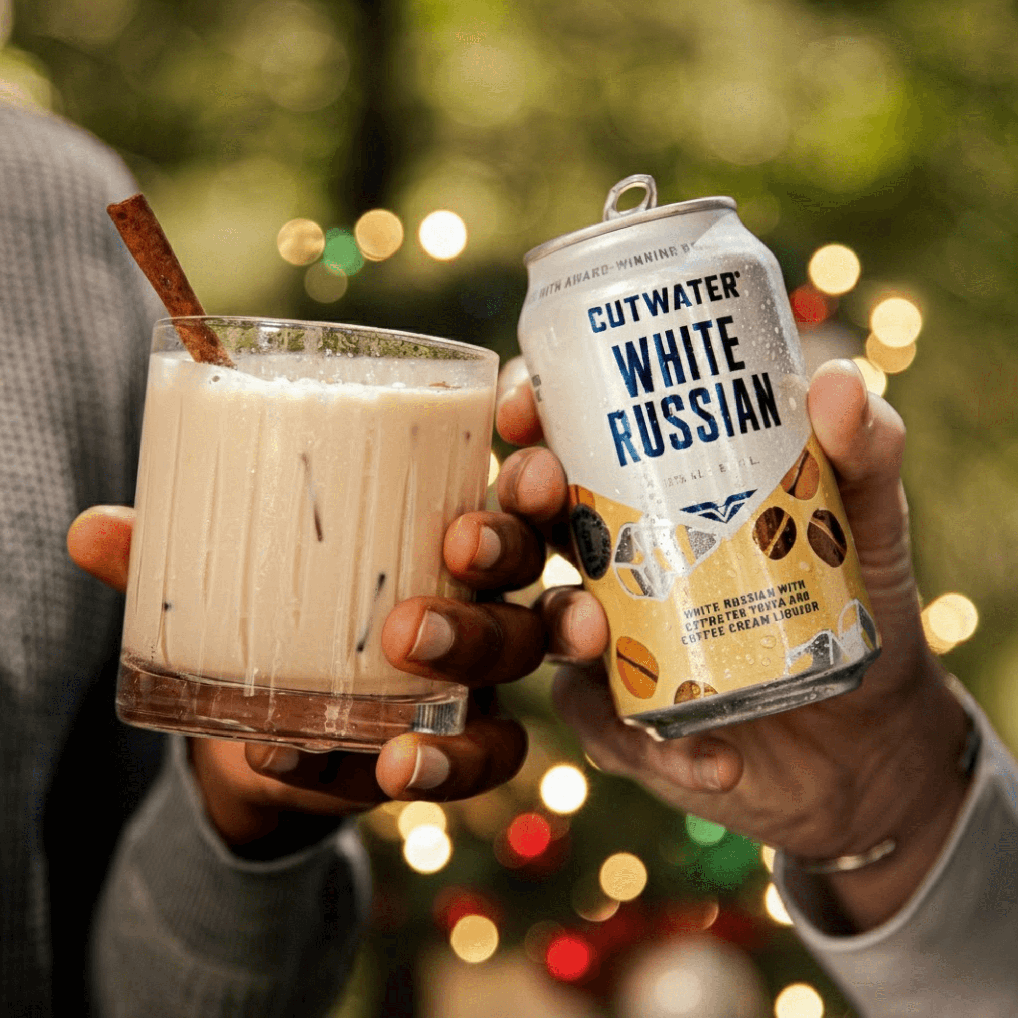 Cutwater Holiday Cocktails: Festive Flavors for Every Celebration Hands holding a Cutwater White Russian can and a glass of iced cocktail with Christmas lights in the background.