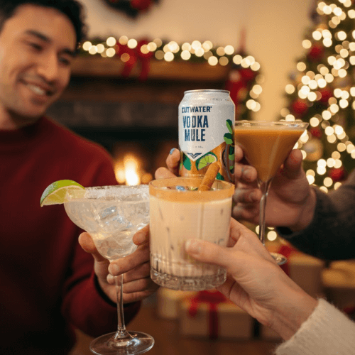 Friends toasting with holiday cocktails and a Cutwater Vodka Mule can in front of a Christmas tree with warm festive lighting.