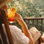 A person wearing a sun hat, sitting in a wooden rocking chair on a porch and holding a layered Texas Sunrise cocktail during golden hour.