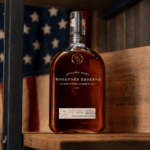 Inside Zipps' Veterans Giving Program: How Your Purchase Supports Texas Communities Woodford Reserve bourbon bottle displayed on a rustic wooden shelf with a softly blurred American flag in the background.