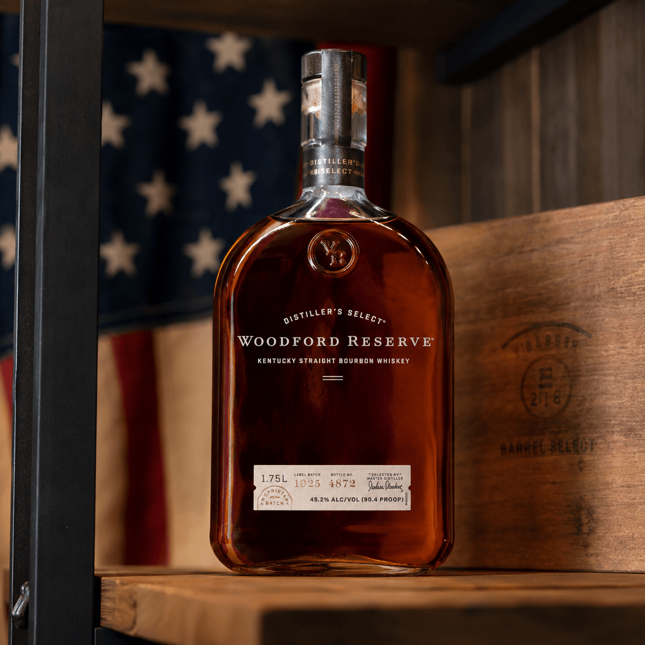 Woodford Reserve bourbon bottle displayed on a rustic wooden shelf with a softly blurred American flag in the background.